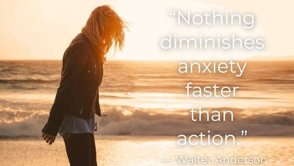 Blonde woman in a black jacket on the beach looking down at her shoes with quote: “Nothing diminishes anxiety faster than action.” — Walter Anderson