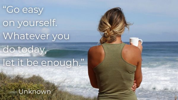 Woman with braided hair standing facing the ocean holding a coffee cup with quote: “Go easy on yourself. Whatever you do today, let it be enough.” — Unknown