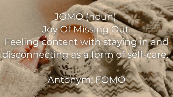A hand holding a mug filled with coffee resting on a fuzzy blanket with quote: 	JOMO (noun) Joy Of Missing Out.  Feeling content with staying in and disconnecting as a form of self-care.  Antonym:  FOMO
