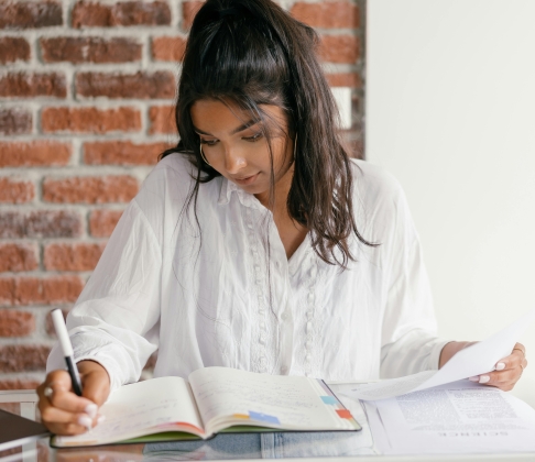 Dauntless Counseling Anxiety Therapy for Women - Image of a young woman with black hair pulled back in a pony tail sitting and writing in a notebook with a red brick wall behind her