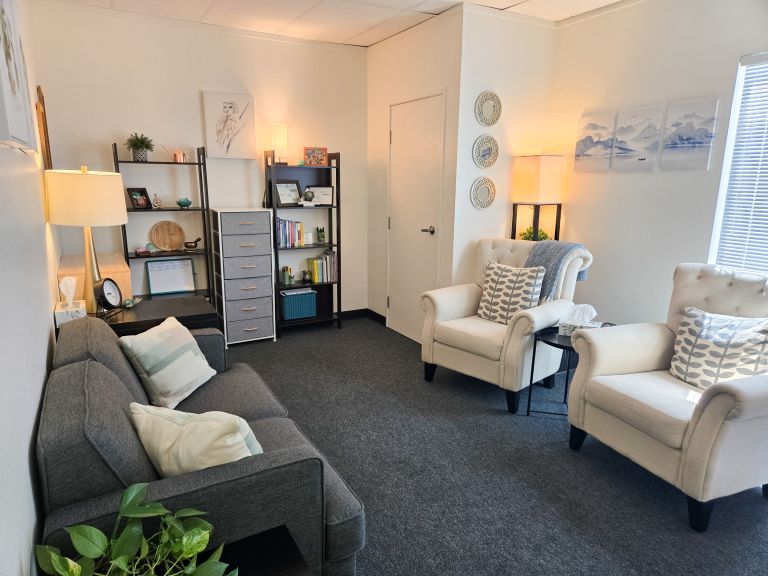 Dauntless Counseling Location - Office with two cream colored arm chairs, two book shelves, a gray couch, gray drawers, and multiple plants and lamps